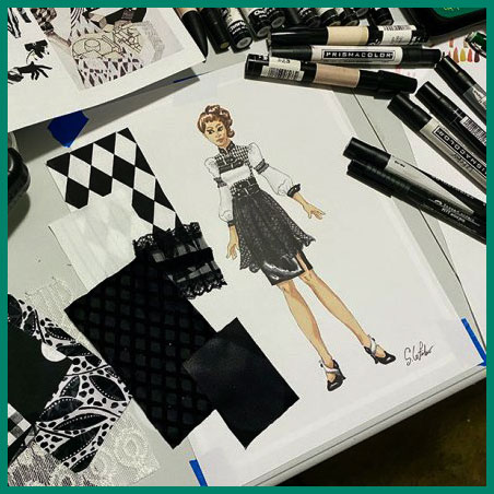 A work table covered with markers, fabric swatches, inspiration images, and a fashion illustration rendered in art markers. The illustration is a modern edgy design.