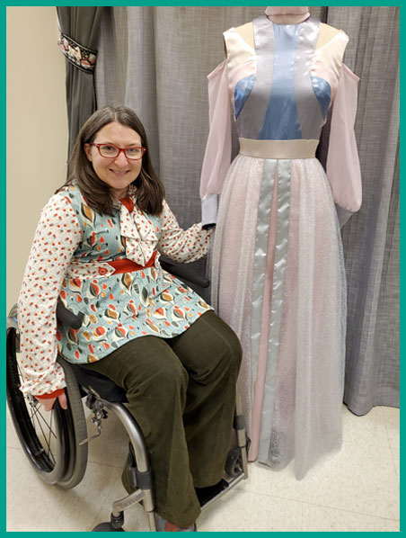 A picture of Sarah next to a dress form wearing a high fashion original dress design. Sarah is seated in a wheelchair, smiling, and wearing a vintage-inspired original design handmade top. On the dress form is a floor length futuristic elegant gown made of satin and chiffon in multiple pastel colors.