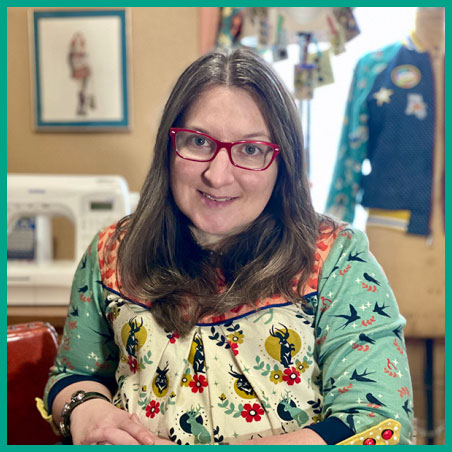 A picture of Sarah - a smiling woman seated in the sewing studio wearing a colorful handmade top. Behind her is a sewing machine and a dress form wearing a handmade jacket.